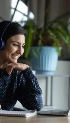 A imagem mostra uma mulher sentada a uma mesa de trabalha, em frente a seu notebook e com fones de ouvidos. Ela está sorrindo.