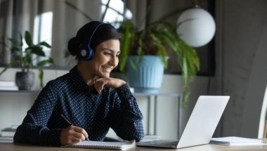 A imagem mostra uma mulher sentada a uma mesa de trabalha, em frente a seu notebook e com fones de ouvidos. Ela está sorrindo.