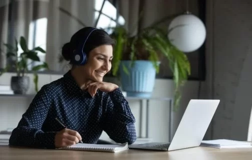 A imagem mostra uma mulher sentada a uma mesa de trabalha, em frente a seu notebook e com fones de ouvidos. Ela está sorrindo.
