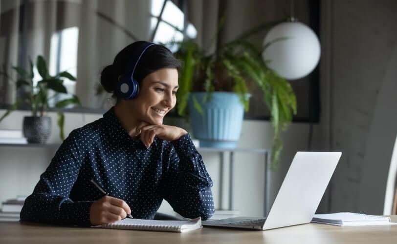 A imagem mostra uma mulher sentada a uma mesa de trabalha, em frente a seu notebook e com fones de ouvidos. Ela está sorrindo.