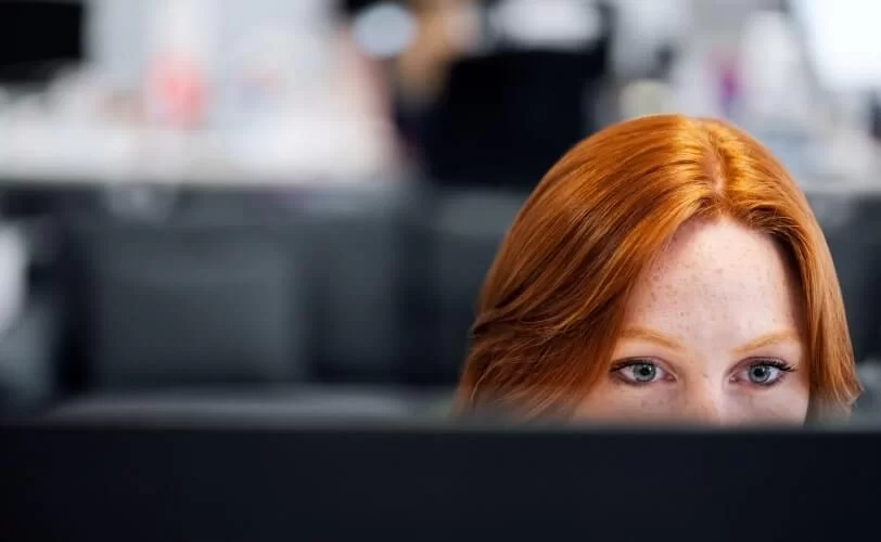 Na imagem vemos uma mulher ruiva atrás de uma tela de computador. 