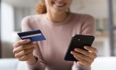 a imagem mostra uma mulher segurando um cartão de crédito em uma mão e o celular em outra. Ela está sorrindo.