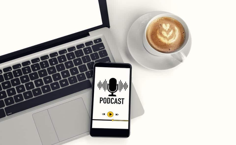 Na imagem há um notebook, xícara de café e um smartphone escrito 'podcast' na tela. 