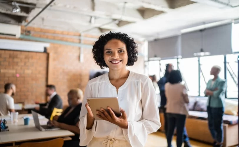 Mulher em um escritório com diversas pessoas ao fundo. Ela está segurando um tablet e sorrindo para a foto. 