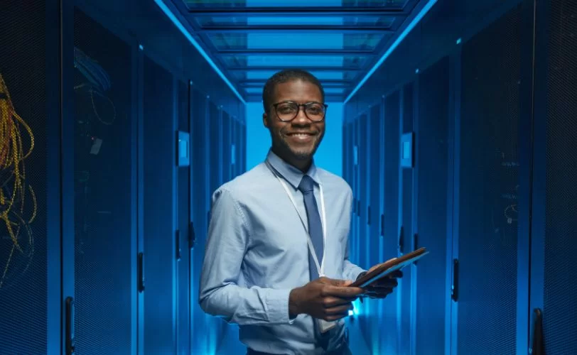 Homem em um data center, sorrindo e segurando um tablet. Ele veste camisa azul clara e gravata azul escura. O ambiente é iluminado por luzes azuis e cercado por racks de servidores pretos. Cabos amarelos são visíveis à esquerda, indicando uma infraestrutura de rede organizada.