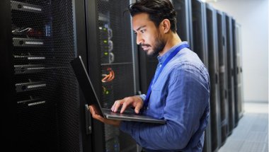 Homem com laptop em frente a um servidor para manutenção, segurança cibernética e verificação de internet.
