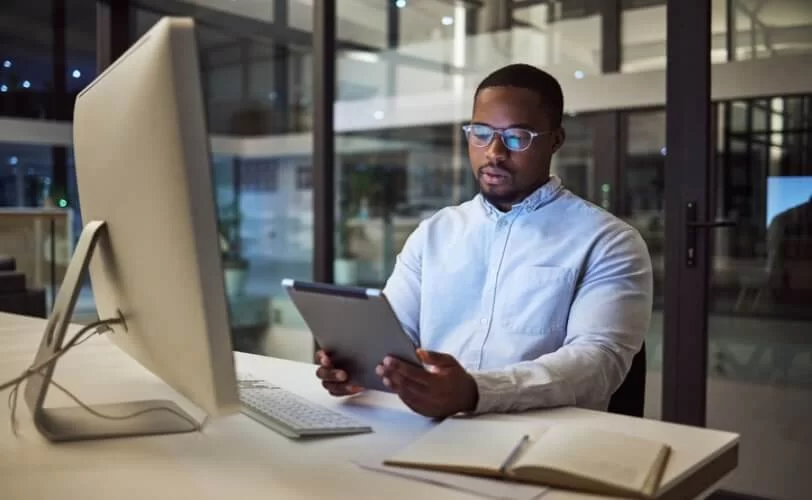 rapaz sentado em uma mesa de escritório, olhando um tablet e tem um computador a sua frente. 