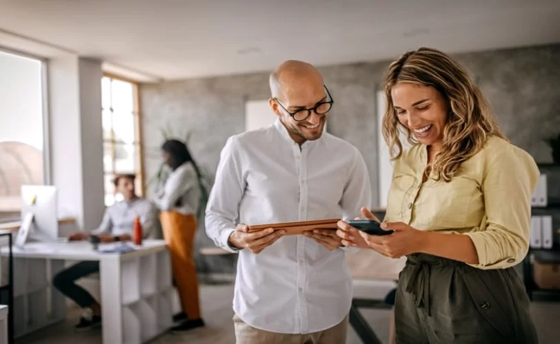 Homem e mulher lado a lado. Ele está com um tablet na mão. Ela está com um smartphone. Eles estão conversando e rindo. 