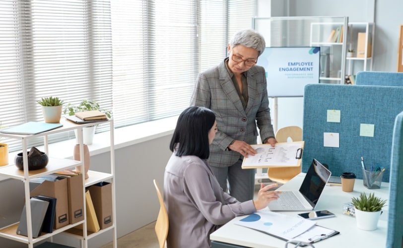 Duas mulheres em um escritório criativo. Uma está de pé mostrando gráficos em uma prancheta para outra mulher que está sentado em frente a um computador. 