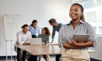 A imagem mostra uma sala de escritório. A frente está uma moça sorrindo e no fundo quatro pessoas conversando.