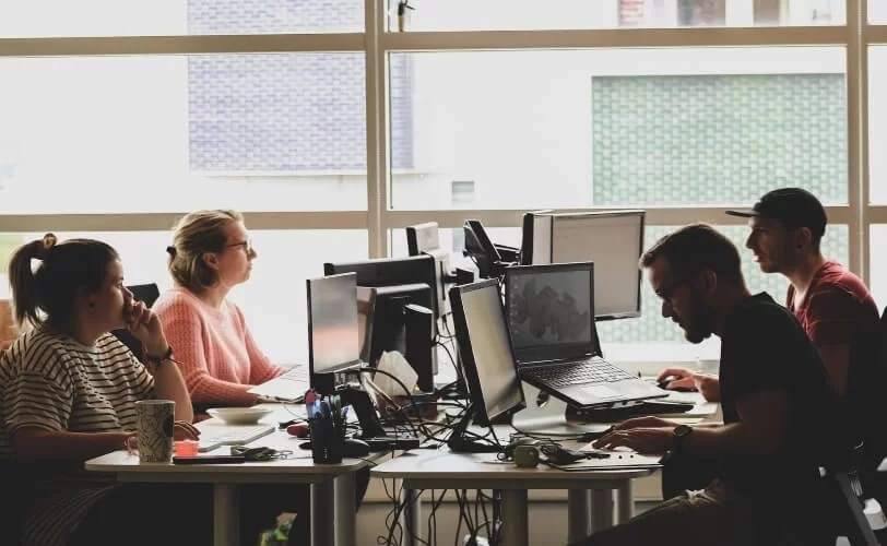 quatro pessoas sentadas em volta de uma mesa de um escritório. Há computadores e várias telas em cima da mesa. 