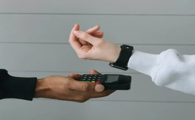 Na imagem aparece a mão de um homem segurando uma maquininha de pagamento e outro homem aproximando o seu smartwatch do aparelho. 