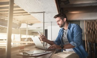 Homem sentado em frente a uma janela com seu notebook e smartphone a mão.