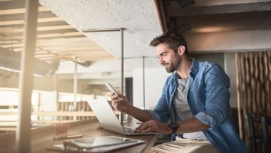 Homem sentado em frente a uma janela com seu notebook e smartphone a mão.