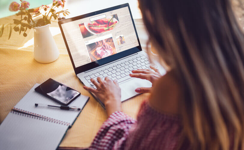 Uma mulher está de costas para a imagem. Ela veste roupas casuais e está sentada em frente a uma mesa, escrevendo um blog post em seu notebook. Ao lado dela estão um caderno, uma caneta e um smartphone.