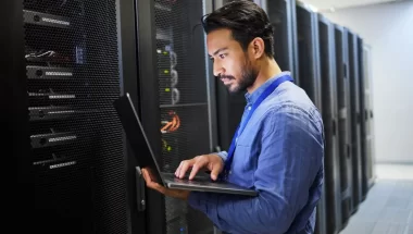 A imagem mostra um homem dentro da sala de servidores de uma empresa. Ele está digitando no teclado de um notebook, possivelmente realizando algumas configurações de servidor proxy.