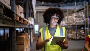 A imagem mostra uma mulher trabalhando no estoque do que parece ser um grande varejo. Ela está digitando algumas informações importantes em um tablet, aplicando conceitos variados, como o que é SKU.