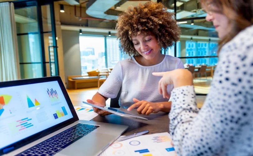 duas mulheres sentadas a uma mesa e ambas estão conversando sobre alguns dashboards que estão na tela de um notebook. 