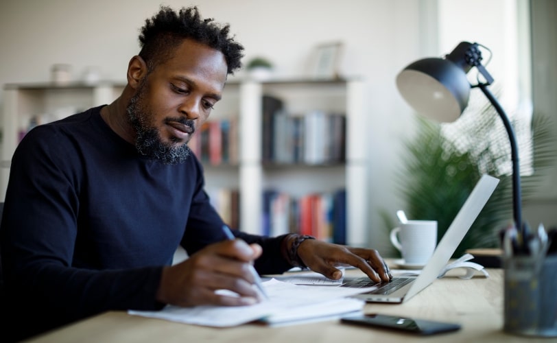 Homem sentado a uma escrivaninha com uma mão sobre um notebook e com a outra mão ele está escrevendo em caderno. 