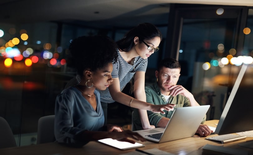 A imagem mostra três pessoas em um escritório olhando para a tela de um notebook. A foto faz parte do conteúdo sobre Google Analytics e sua importância no Marketing Digital. 