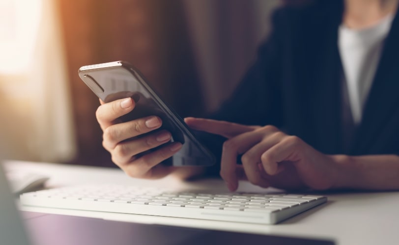 Mulher usando um smartphone e próximo a suas mãos há um teclado de computador. A imagem compõe o conteúdo sobre GA e marketing digital. 