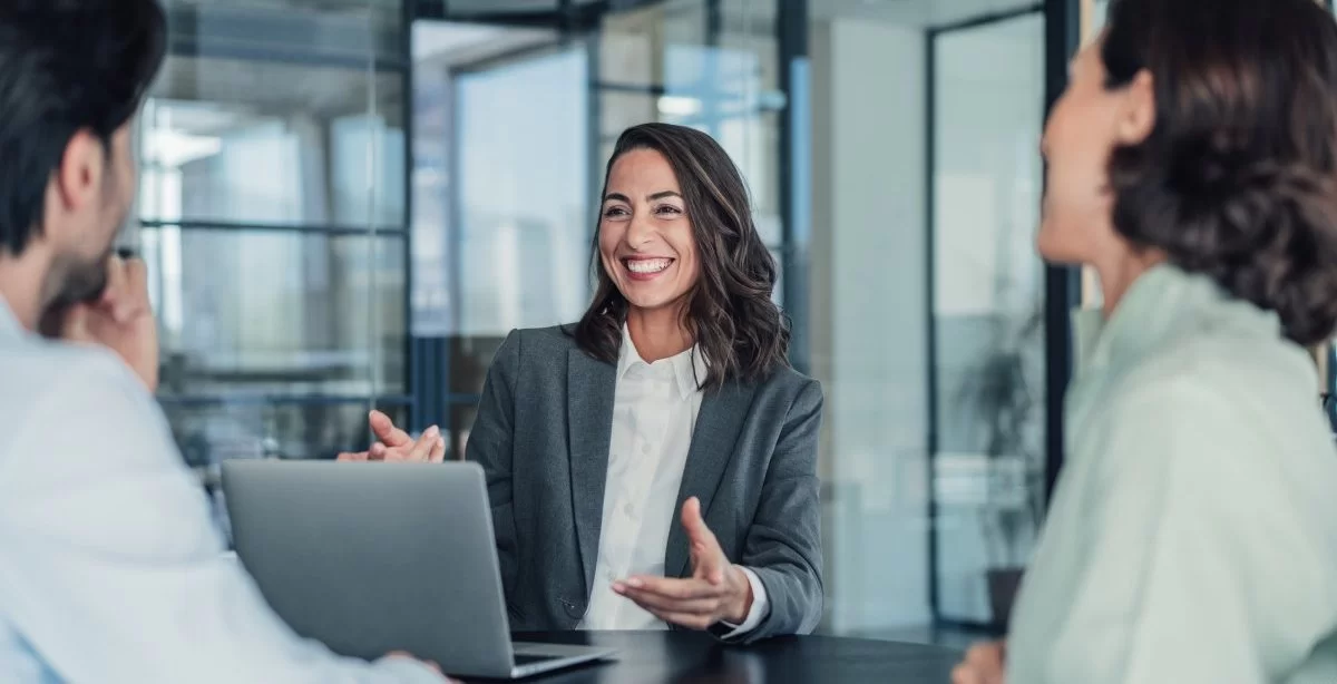 A imagem mostra uma mulher conversando com duas pessoas. Ela está em um escritório, possivelmente aplicando conceitos de marketing pessoal para destacar as habilidades dela.