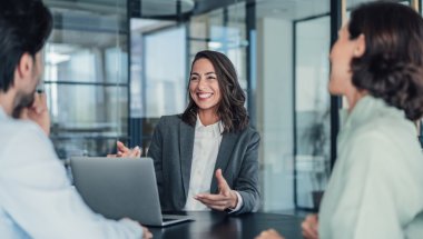 A imagem mostra uma mulher conversando com duas pessoas. Ela está em um escritório, possivelmente aplicando conceitos de marketing pessoal para destacar as habilidades dela.