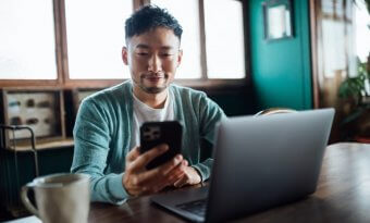 Homem desenvolvedor sentado em uma escrivaninha com um notebook a sua frente e olhando seu smartphone