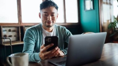 Homem desenvolvedor sentado em uma escrivaninha com um notebook a sua frente e olhando seu smartphone