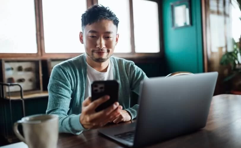 Homem desenvolvedor sentado em uma escrivaninha com um notebook a sua frente e olhando seu smartphone