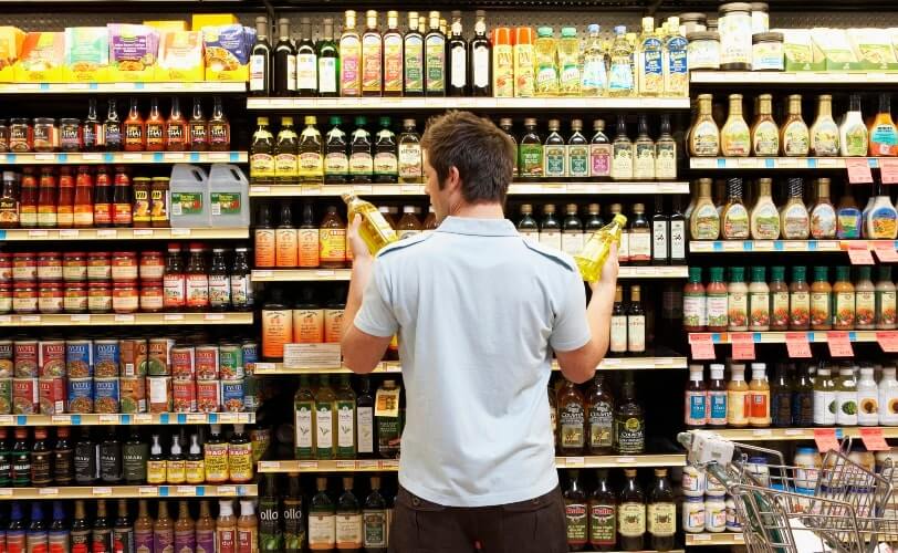 Homem em frente a uma prateleira de óleos de cozinha no supermercado. 