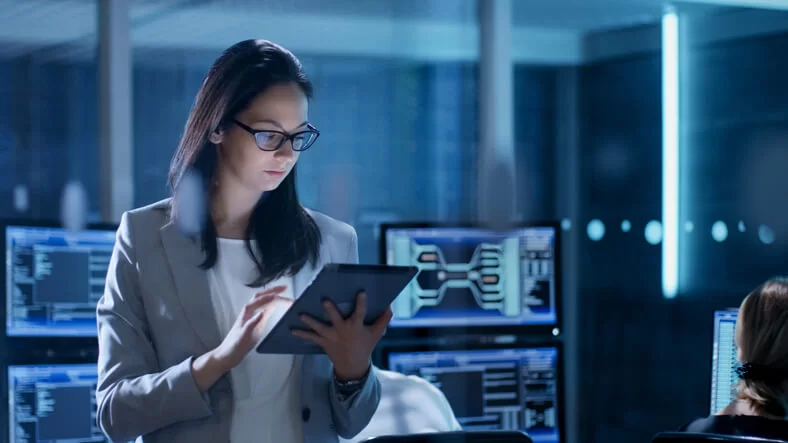 Uma mulher em um escritório moderno trabalha em um tablet. Vários monitores de computador exibindo dados estão em segundo plano. Ela tem a pele branca, os cabelos pretos, usa óculos de grau e veste uma camiseta branca e um terno cinza.