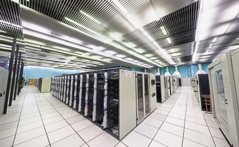 A imagem retrata um data center com fileiras de servidores organizados em racks dentro de um ambiente controlado. O corredor entre os servidores é bem iluminado, com piso elevado para ventilação e gerenciamento de cabos. O design reforça a infraestrutura de TI moderna utilizada para armazenamento de dados, Hospedagem Dedicada e em nuvem, e processamento de alto desempenho. 