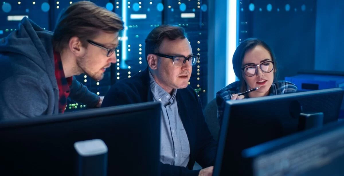 A imagem mostra dois homens e uma mulher conversando em uma sala com servidores. Um deles está digitando em um computador, enquanto todos avaliam o que está na tela, e possivelmente estão falando sobre conceitos aplicados de lógica de programação.