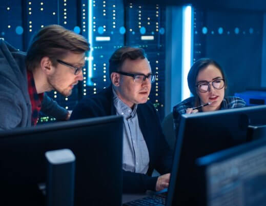 A imagem mostra dois homens e uma mulher conversando em uma sala com servidores. Um deles está digitando em um computador, enquanto todos avaliam o que está na tela, e possivelmente estão falando sobre conceitos aplicados de lógica de programação.