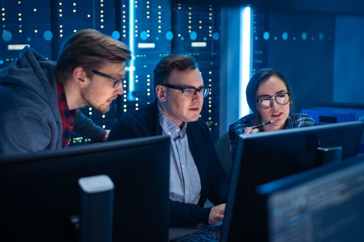 A imagem mostra dois homens e uma mulher conversando em uma sala com servidores. Um deles está digitando em um computador, enquanto todos avaliam o que está na tela, e possivelmente estão falando sobre conceitos aplicados de lógica de programação.
