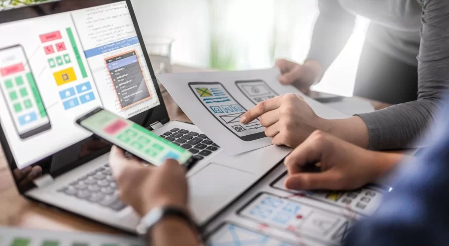 Profissionais utilizando laptops e smartphones em um ambiente de trabalho colaborativo e produtivo. Juntos, eles trocam ideias sobre o layout de um site que aparece na tela. Um deles segura um papel que mostra o planejamento inicial. Os rostos deles não aparecem. 