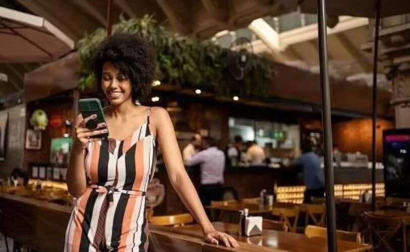 Na imagem há uma moça sorridente em um restaurante, olhando para o celular e sorrindo. A foto faz parte do conteúdo sobre estratégia no youtube.