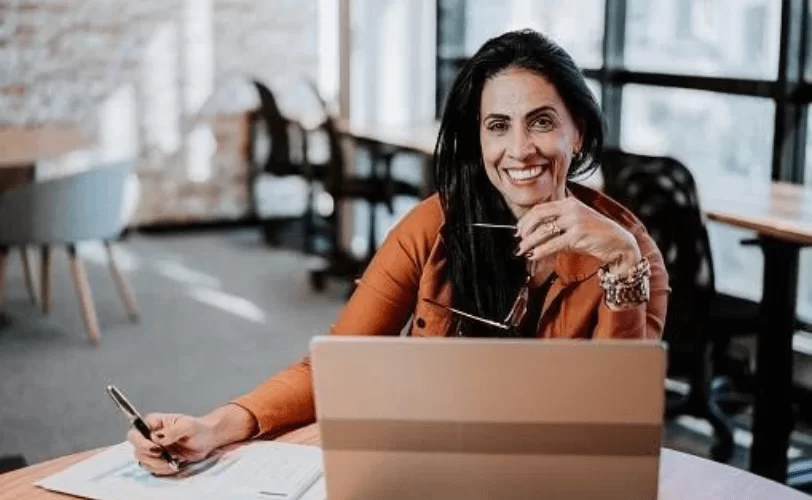 Mulher programadora sorrindo para foto. Ela está em um ambiente de escritório e em frente a um notebook. A imagem faz referência ao IDE, Ambiente de Desenvolvimento Integrado.  
