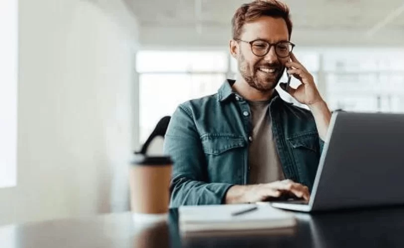  Homem desenvolvedor em frente ao seu notebook e falando com alguém pelo celular sobre ambiente de desenvolvimento integrado.  
