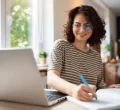 Mulher sorridente em frente a um notebook e escrevendo um plano de metas em um caderno.