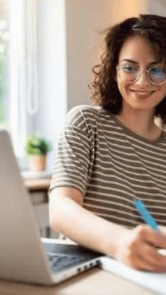 Mulher sorridente em frente a um notebook e escrevendo um plano de metas em um caderno.