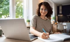 Mulher sorridente em frente a um notebook e escrevendo um plano de metas em um caderno.