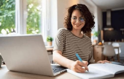 Mulher sorridente em frente a um notebook e escrevendo um plano de metas em um caderno.