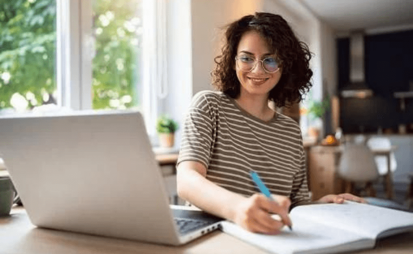 Mulher sorridente em frente a um notebook e escrevendo um plano de metas em um caderno.