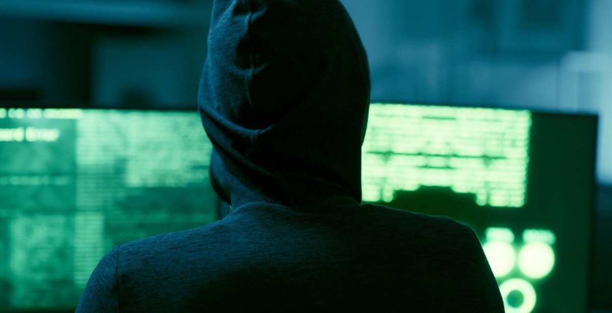 A imagem mostra uma pessoa encapuzada em uma sala com computadores. As telas emitem luzes verdes, com diversos códigos aparecendo em cada uma delas.