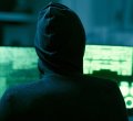 A imagem mostra uma pessoa encapuzada em uma sala com computadores. As telas emitem luzes verdes, com diversos códigos aparecendo em cada uma delas.