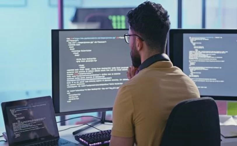 Um rapaz sentado em frente a três telas de computador com códigos abertos no visor. A imagem compõe o conteúdo sobre importância do versionamento de software. 
