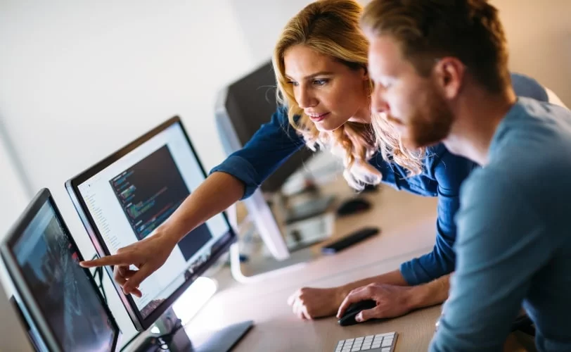 A imagem mostra uma mulher e homem desenvolvedores sentados em frente a seus computadores, conversando sobre versionamento de software. 