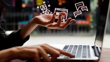 A imagem mostra uma mulher sentada em frente a um notebook e acima da suas mãos há hologramas de ícones da internet.